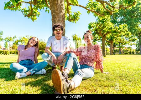 Tre giovani amici diversi che riposano sul campo verde del parco sorridendo guardando la fotocamera per un ritratto. Gen z studenti trascorrere il tempo in natura a rompere c Foto Stock