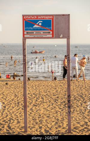 Cartello che vieta di nuotare a 2 chilometri di distanza sulla spiaggia del Golfo finlandese, scritto in russo o cirillico, con persone che nuotano in acqua Foto Stock