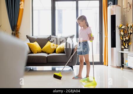 Piccola ragazza bionda caucasica con capelli biondi sette anni piano di pulizia in soggiorno. Interni moderni, vita domestica. Foto Stock