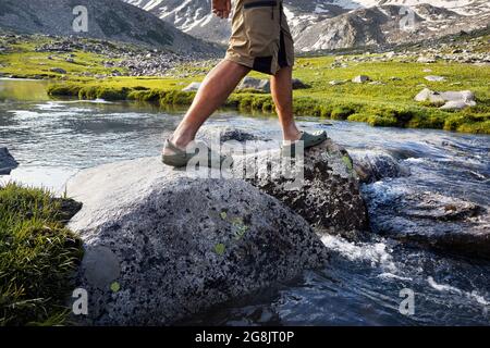 Uomo escursionistico che attraversa il fiume in scarpe verdi da pietre in equilibrio su caduto in Kazakistan Foto Stock