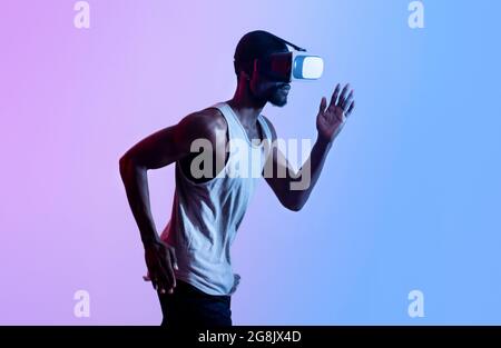 Vista laterale di un atleta nero millenario con visore VR che funziona con illuminazione al neon. Tecnologie contemporanee nello sport Foto Stock
