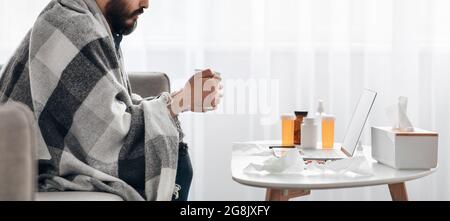 Concetto di febbre. Malato ragazzo si sente malato e bere tè caldo, seduto sul divano e guardando film sul computer portatile, vista laterale, raccolto Foto Stock