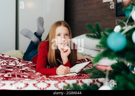 Ragazzina caucasica dai capelli rossi che giace sul letto scrivendo una lettera a Babbo Natale. Foto Stock