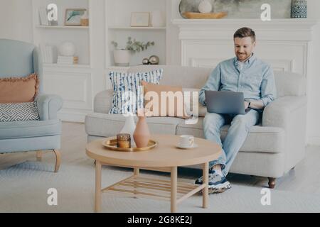 Giovane uomo che lavora come freelance sul computer portatile mentre si siede sul divano Foto Stock