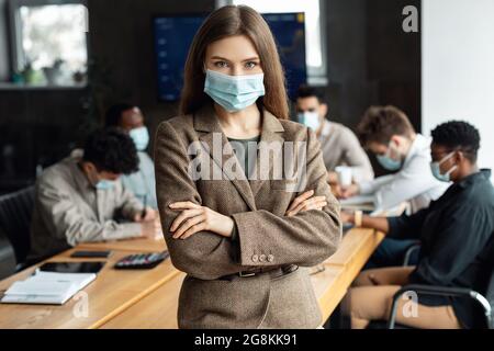Giovane donna d'affari in maschera seduta sulla scrivania e in posa Foto Stock