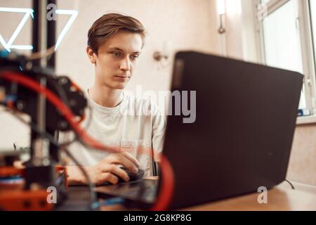 Un giovane uomo concentrato nella creazione di un modello 3D su un computer portatile per la stampa mentre si è seduti a tavola e si lavora a casa Foto Stock