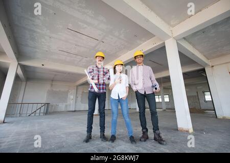 Un team di ingegneri della Diversity, una donna e due uomini, in piedi insieme e sorridendo alla macchina fotografica mentre si lavora in un cantiere di costruzione di edifici interni. Foto Stock