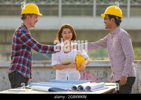 Due ingegneri, asiatici e caucasici che scuotono le mani dopo il lavoro è finito sul tetto del cantiere con una femmina in squadra guardando e sorridendo con h Foto Stock