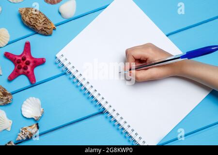 Il blocco note di vuoto e la mano con una penna diverse voci marino nel blu dipinto di sfondo di legno. Mare di oggetti su tavole di legno. Messa a fuoco selettiva. Posto per t Foto Stock