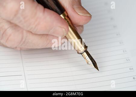 Penna d'inchiostro fontana color oro in mano donna anziana circa per scrivere qualcosa sulla pagina vuota del pianificatore giornaliero, dettaglio closeup Foto Stock