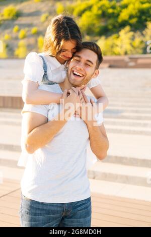 Ritratto verticale di felice giovane coppia che si diverte nel parco cittadino in estate soleggiato giorno, giovane donna appesa sulle spalle di amante ridente. Foto Stock