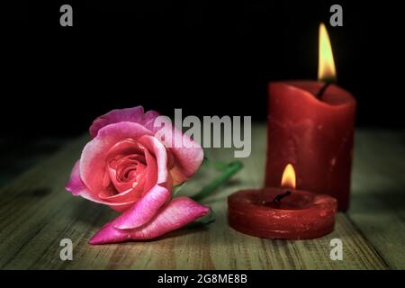 Fioritura di una rosa che giace su un tavolo di legno accanto ad un paio di candele rosse accese Foto Stock