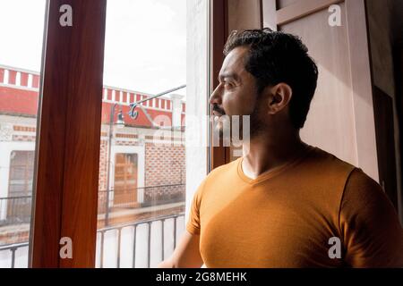 Bell'uomo ispanico con un bel portamonete con una t-shirt arancione che guarda fuori dalla finestra Foto Stock