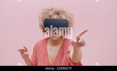 Ragazza che indossa occhiali di realtà virtuale guardando un film in 3D. Sentirsi stupiti e puntare allo schermo virtuale. La ragazza si perde nella realtà virtuale. Isolato su sfondo rosa studio. Foto Stock