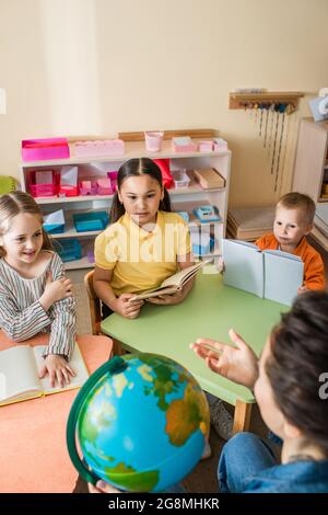 insegnante che punta con la mano vicino al globo davanti ai bambini interracial Foto Stock