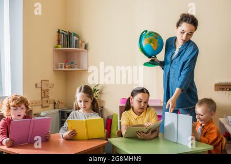 insegnante con il globo che punta al libro vicino i capretti interracial che leggono in classe Foto Stock