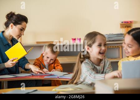 insegnante che indica il taccuino vicino al ragazzo di scrittura mentre le ragazze interrazziali ridono sul primo piano offuscato Foto Stock