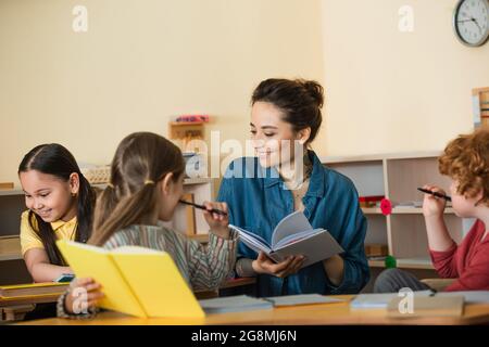 felice insegnante che tiene un libro di testo vicino a bambini multietnici in classe Foto Stock