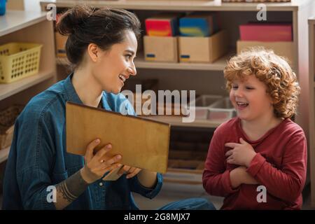 felice insegnante che mostra tavola di legno mentre si gioca a gioco educativo con riccio ragazzo Foto Stock