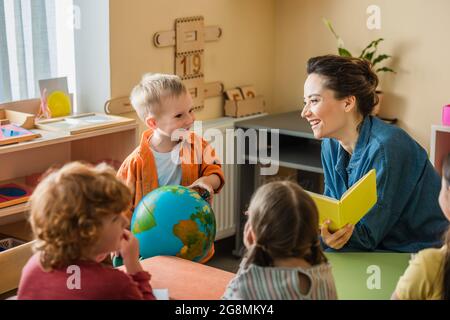 happy boy tenere il globo vicino a sorridente insegnante e bambini multietnici Foto Stock