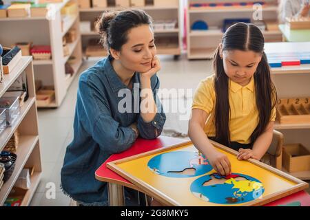 giovane insegnante che guarda il bambino asiatico che combina puzzle mappa della terra nella scuola di montessori Foto Stock