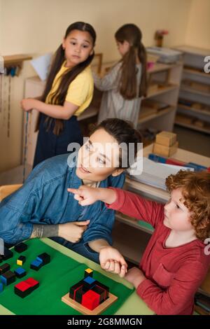 ragazzo che punta con il dito mentre si gioca a cubetti di legno gioco vicino insegnante e ragazze interracial offuscate Foto Stock