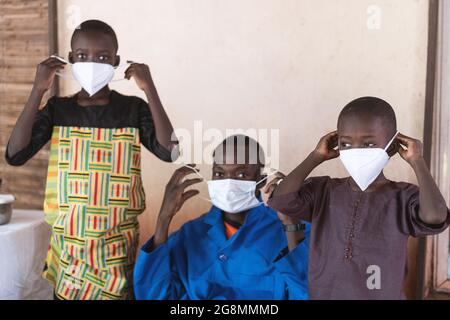 In questa immagine un operatore sanitario africano seduto in un cappotto di laboratorio blu sta mostrando come indossare e montare una maschera antivirale protettiva con il supporto di due Foto Stock