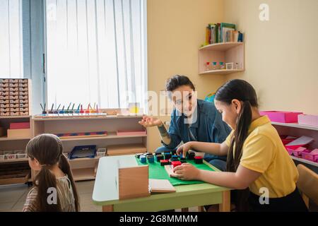 insegnante sorridente gesturing mentre si gioca a cubi multicolore gioco con ragazza asiatica Foto Stock