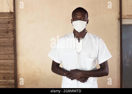 Questa foto mostra un giovane medico africano sicuro con uno stetoscopio piegando le mani e guardando direttamente nella macchina fotografica indossando una maschera facciale e un Foto Stock