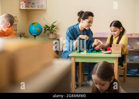 insegnante che punta a cubi multicolore mentre gioca con ragazza asiatica vicino al bambino in primo piano bllurred Foto Stock