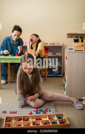 bambino sul pavimento vicino lettere di legno e insegnante gioco con ragazza asiatica su sfondo sfocato Foto Stock