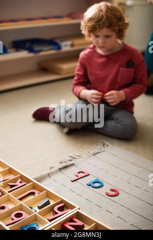 bambino offuscato seduto sul pavimento vicino a lettere di legno nella scuola di montessori Foto Stock