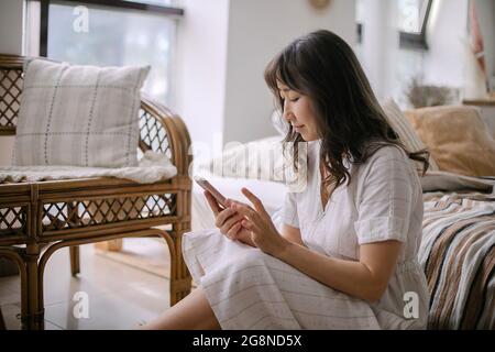 Giovane bella donna asiatica in abito estivo bianco con smartphone mentre si rilassa a casa Foto Stock
