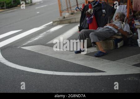 Uomo senza tetto che dorme nella striscia mediana con i suoi vestiti e altri beni di famiglia sotto la strada della città terra ponte elevato, in Giappone, giorno nuvoloso Foto Stock