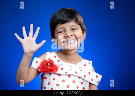 Carino capretto della ragazza che dice hi anf come sono guardando la macchina fotografica su sfondo blu di colore - concetto di saluto del bambino. Foto Stock