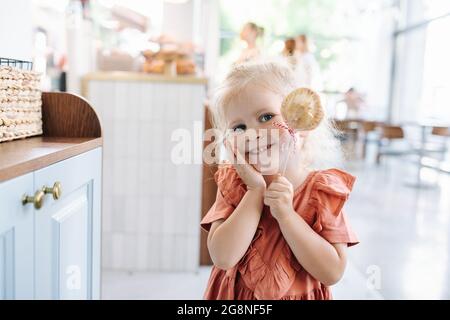 Adorabile bambina bionda che tiene il biscotto su un bastone, sorridendo alla macchina fotografica, giocando carino. Foto Stock