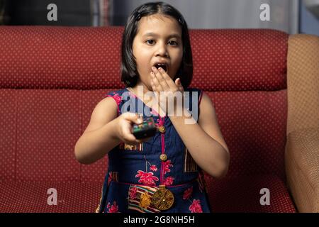 Una bambina indiana urla mentre guardi la televisione con telecomando in mano e seduto sul divano Foto Stock