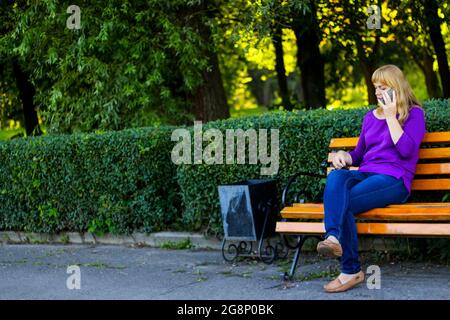 Defocus bionda caucasica donna che parla, parlando al telefono esterno, all'aperto. donna di 40 anni in blusa viola in panchina. Adulto femmina usi Foto Stock