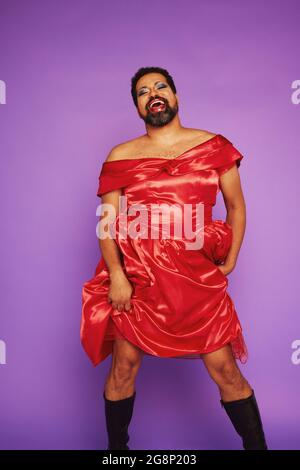 Uomo gay che indossa trucco e abito femminile. Queer che dà una performance di canto su sfondo viola. Foto Stock