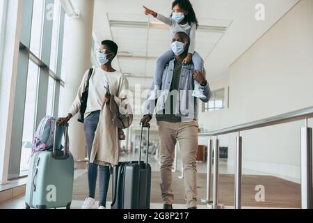 Padre africano con la figlia giovane a spalla camminando lungo la moglie con i bagagli al terminal dell'aeroporto. Famiglia in pandemia con bagagli all'aeroporto corrido Foto Stock