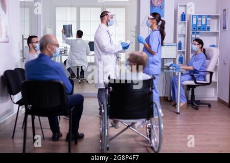 Team medico con visiera e maschera di protezione che discute in piedi in sala d'attesa della clinica medica prima dell'esame dei pazienti. Anziani in visita medica durante la cofid 19 pandemia globale Foto Stock