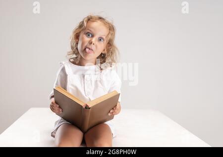 Studio shot di piccola cute caucasica ragazza con libro mettendo fuori la sua lingua isolato su sfondo bianco studio. Foto Stock