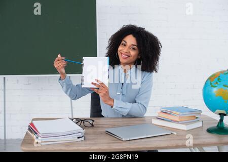 Sorridente insegnante afroamericano che punta al notebook vicino al portatile e al mondo in classe Foto Stock