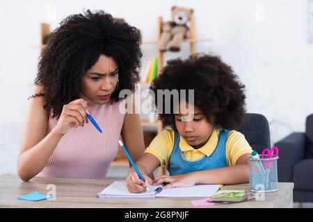 Arrabbiata madre afroamericana indicando con penna vicino figlia che scrive sul notebook a casa Foto Stock