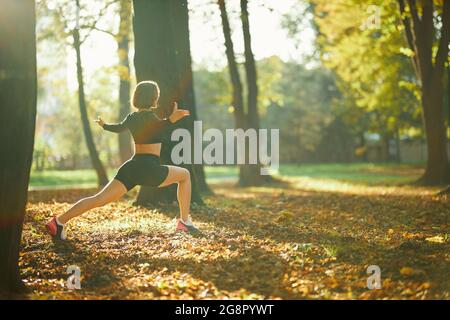 Fitness giovane donna vestita con crop top nero e pantaloncini sportivi che allungano le gambe durante una giornata di sole al parco cittadino. Tempo libero all'aria aperta e stile di vita attivo. Foto Stock