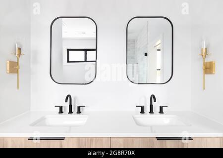 Un bagno splendidamente rinnovato con un armadietto in legno, specchio con cornice nera e rubinetti, e luci dorate montate sulla parete. Foto Stock