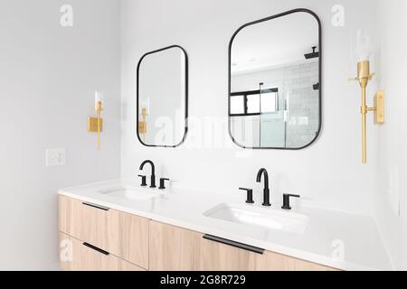 Un bagno splendidamente rinnovato con un armadietto in legno, specchio con cornice nera e rubinetti, e luci dorate montate sulla parete. Foto Stock