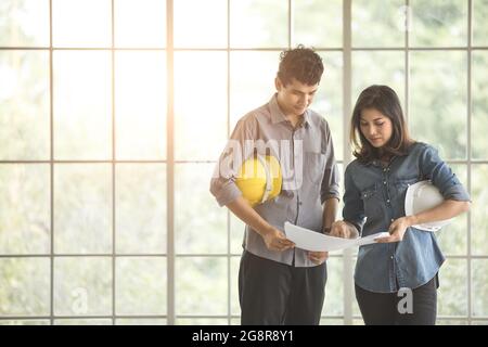 Due ingegneri asiatici maschi e femmine che tengono in piedi caschi di sicurezza bianchi e gialli e parlano insieme. Cornice in vetro sullo sfondo Foto Stock