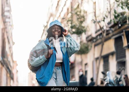 Giovane donna africana fresca con trecce sorridenti e a piedi in città. Indossa abiti casual e un cappello. Foto Stock