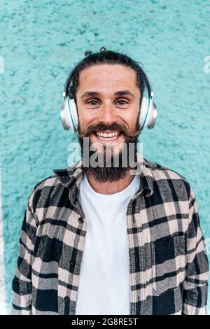 Uomo allegro con capelli lunghi e barba grande sorridente e guardando la macchina fotografica su sfondo blu. Sta ascoltando musica. Foto Stock
